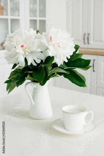 White peonies in a vase and a cup of milk on a white background in the interior. Breakfast. Composition.