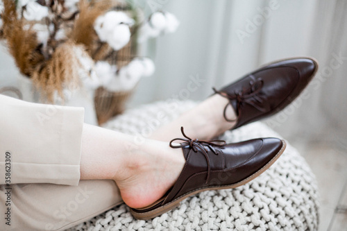 fashionable women's shoes, feet in the interior, classic women’s flat shoes, brown leather shoes, mules and loafers, knitted blankets and blankets on the background, wooden background, cotton flowers,