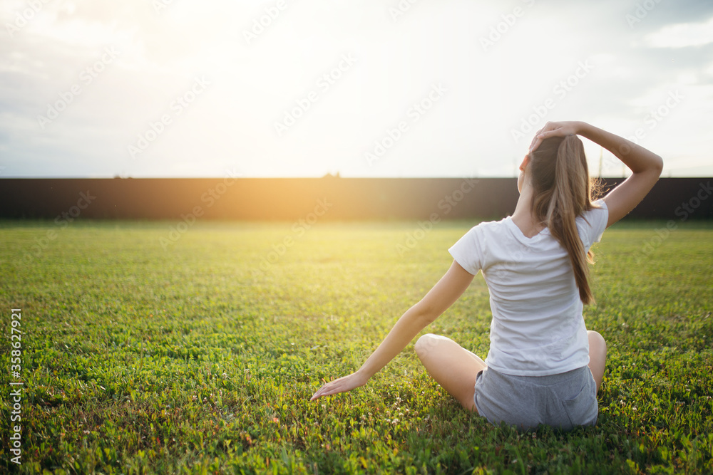 The view from the back of a beautiful girl who is sitting on a green lawn and doing exercises on her neck while sitting in a lotus position. The view from the back of a beautiful girl who is sitting on a green lawn and doing exercises on her neck while sitting in a lotus position.