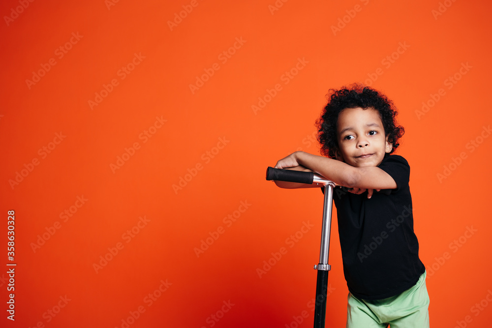 African american boy in black t-shirt leaning his scooter on his hands and head on an orange background in studio