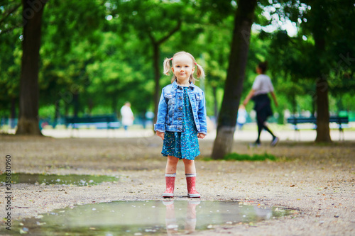 Child wearing red rain boots and jumping in puddle on a summer day. Adorable toddler girl having fun with water and mud in park on a rainy day. Outdoor activities for kids © Ekaterina Pokrovsky