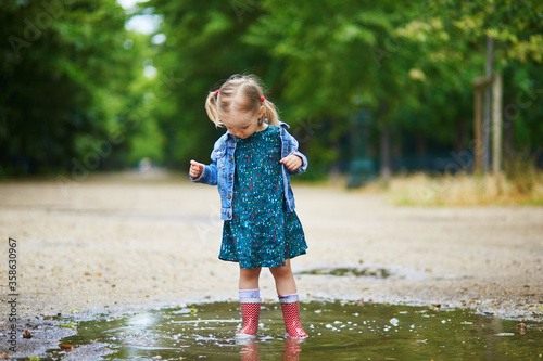Child wearing red rain boots and jumping in puddle on a summer day. Adorable toddler girl having fun with water and mud in park on a rainy day. Outdoor activities for kids © Ekaterina Pokrovsky