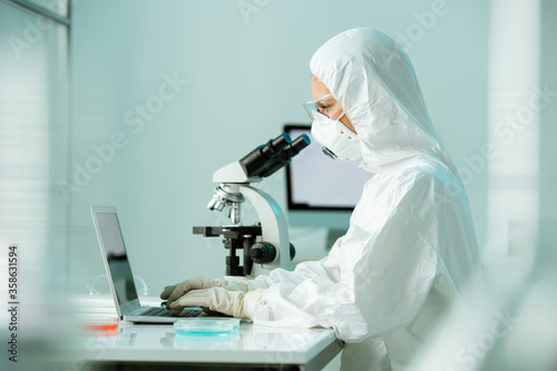Busy lab scientist in protective suit and respiratory mask typing research report about coronavirus infection in laboratory