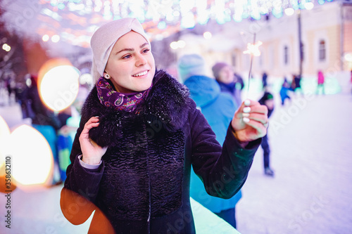 Happy young woman with winter clothes smiles lights sparklers, background evening city illumination. Christmas concept