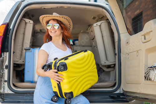 Happy red-haired woman in a cowboy hat and sunglasses going on a car trip around the country. Girl sitting in the trunk with a yellow suitcase ready for summer vacation. Independent travel.