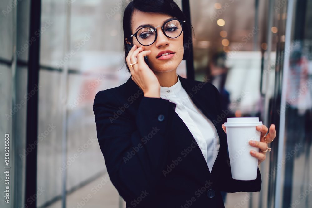 Serious corporate manager talking on phone during coffee break outdoors holding takeaway cup.Businesswoman in formal wear and eyewear enjoying rest having mobile conversation with colleague