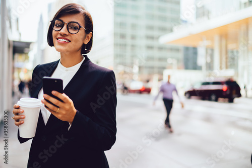 Half length portrait of cheerful brunette corporate boss in optical spectacles enjoying time for coffee break in business district, happy smiling woman in formal wear holding modern smartphone device
