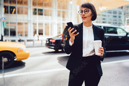 Thoughtful female economist in trendy eyeglasses holding mock up coffee to go cup and looking away standing on street.Trader in elegant formal wear strolling in downtown with telephone in hand