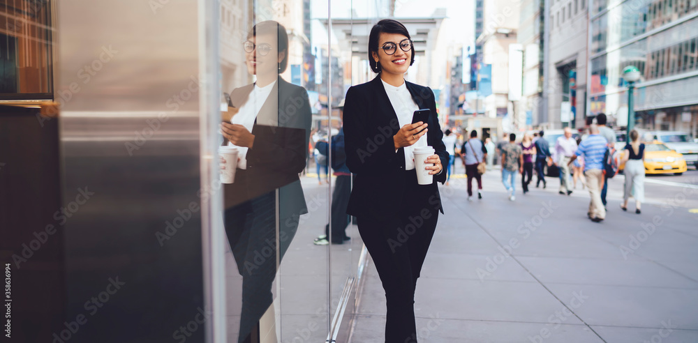 Full length portrait of successful business woman dressed in elegant black suit standing on urban setting in New York and enjoying work break for coffee and time for communicate via cellular phone