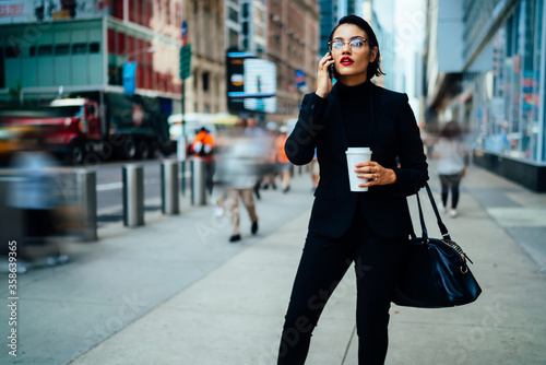 Serious confident business woman in elegant black suit and optical eyewear calling to partner for discussing time of meeting in financial district using roaming connection on modern mobile phone