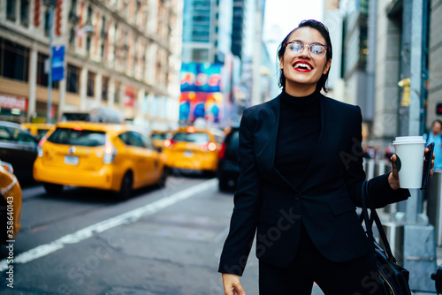 Canvas Print Cheerful female entrepreneur in elegant wear waiting for taxi standing on road with coffee to go, successful businesswoman calling yellow cab in good mood searching transport getting to office