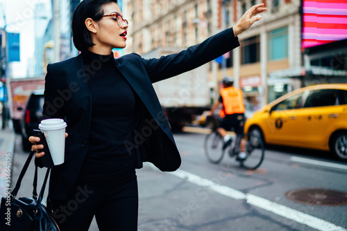 Valokuva Attractive businesswoman with red lips raising had to call yellow cab on new york avenue, confident trendy dressed female manager hailing on road catching taxi holding coffee to go getting to work