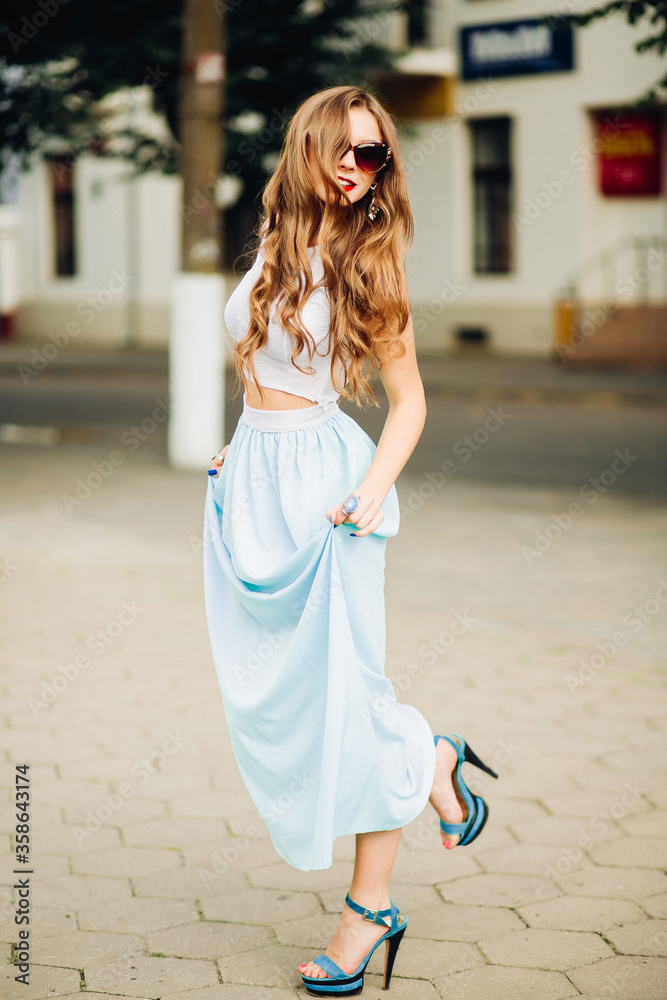 Fashion and stylish girl with long wavy hair turned away smiling and holding long skirt by hands. Woman in sunglasses posing and looking over shoulder, wearing in top and long blue skirt. Summertime.
