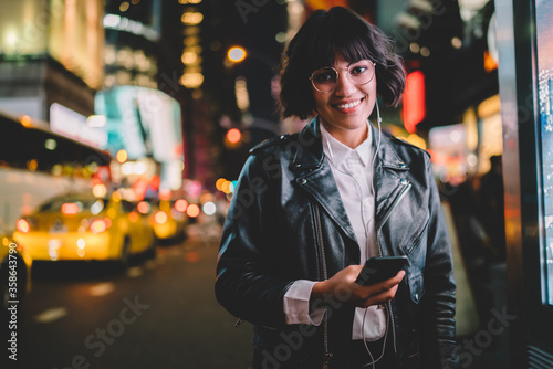 Half length portrait of cheerful millennial girl in fashionable apparel listening positive media record and smiling at camera, happy teenage woman in electronic headphones using cellular device