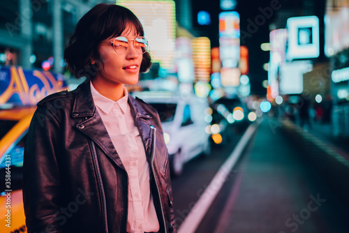 Trendy dressed hipster girl in eyewear with night city light reflection fascinated with beautiful illumination.Gorgeous female in leather jacket wondering while standing on urban background at evening