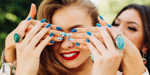 Two beautiful women playing and brunette holding hands and hiding face to her blonde girlfriend. Female with trendy stylish manicure in blue and green tones, fashion accessories, jewelry,big earrings.