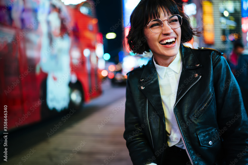 Naklejka premium Pretty positive young woman with short haircut laughing in urban setting with night lights enjoying leisure time in New York.Happy hipster in stylish wear and eyewear having fun on megapolis street