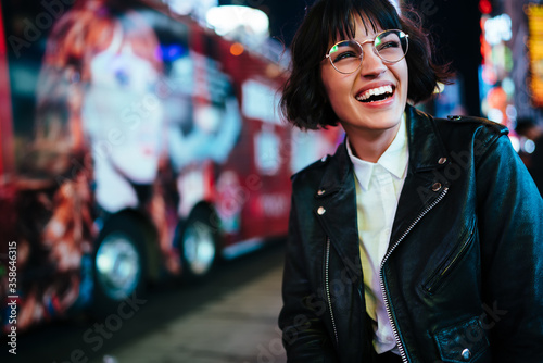 Cheerful hipster girl in design spectacles laughing feeling carefree on free time walking in downtown with night lights.positive young woman in stylish wear having fun in evening megalopolis