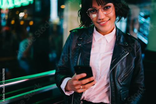 Cropped portrait of cheerful pretty blogger in spectacles dressed in stylish leather jacket smiling at camera while share media in social networkson smartphone via internet 