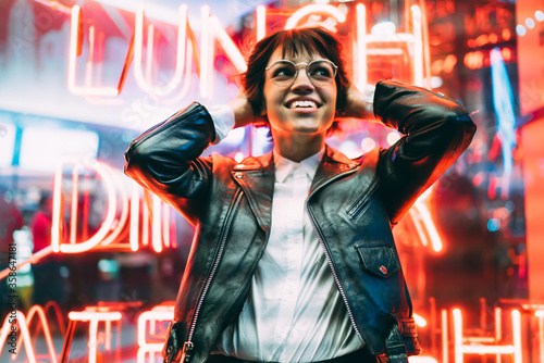 Cheerful hipster girl in trendy apparel excited with night lights in New York City, beautiful brunette woman in spectacles happy about spending evening free time in downtown with neon illumination.