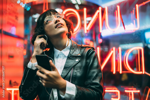 Excited female lover of music dressed in stylish leather jacket listening songs online in earphones connected to smartphone while enjoying night lights and neon illumination in New York City