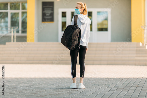 girl in a medical protective mask, standing with a briefcase against the background of a building, in the open air. Quarantine, coronavirus