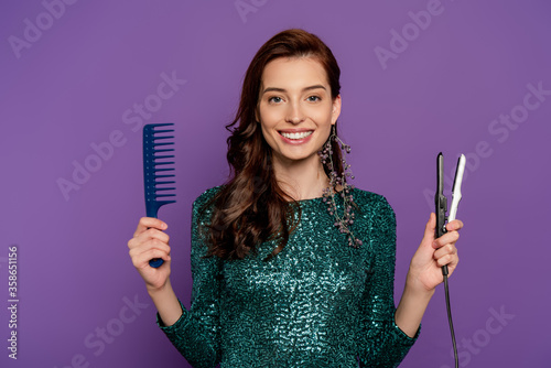 young cheerful woman holding hair straightener and comb isolated on purple