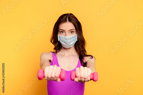 selective focus of sportswoman in medical mask exercising with dumbbells on yellow