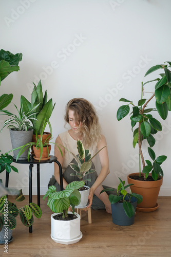 Young blonde girl sits among green plants. Homeliness and flower care in plastic and ceramic pots. Harmony and fresh oxygen in the living room.