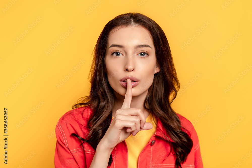 young woman showing hush sign and looking at camera on yellow young woman showing hush sign and looking at camera on yellow