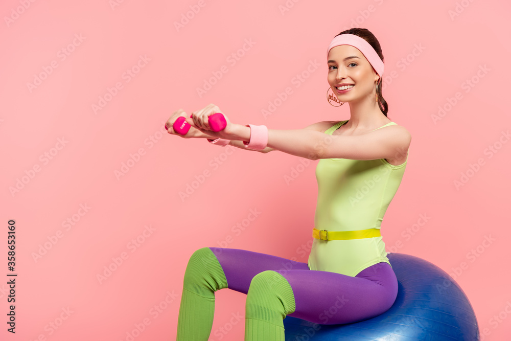 smiling sportswoman with outstretched hands exercising with dumbbells and sitting on fitness ball on pink smiling sportswoman with outstretched hands exercising with dumbbells and sitting on fitness ball on pink