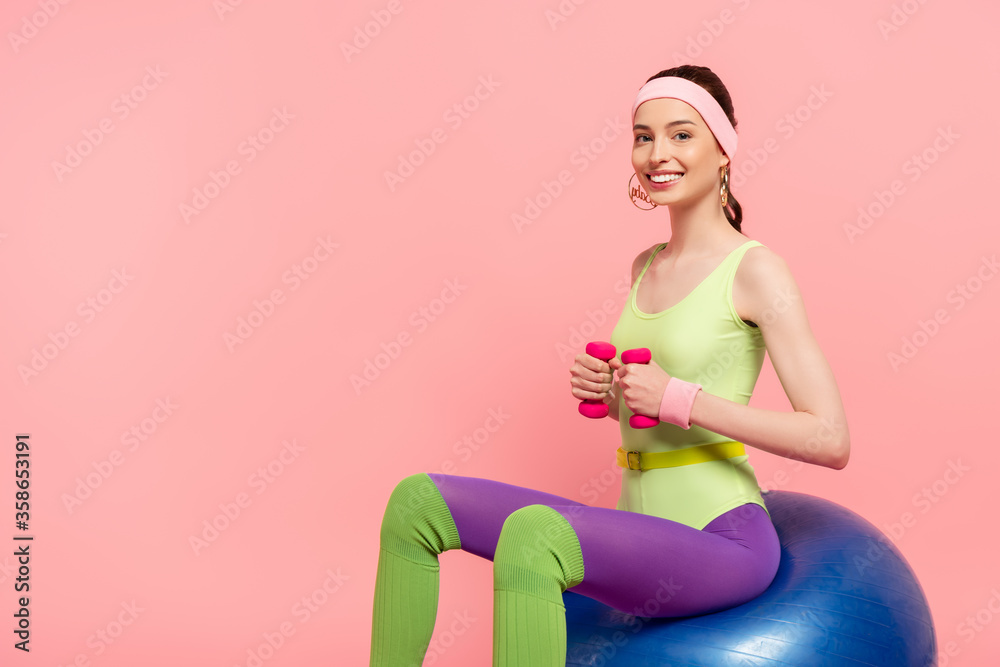 happy woman sitting on fitness ball and exercising with dumbbells isolated on pink happy woman sitting on fitness ball and exercising with dumbbells isolated on pink