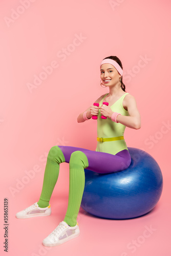positive sportswoman exercising with dumbbells and sitting on fitness ball on pink © LIGHTFIELD STUDIOS