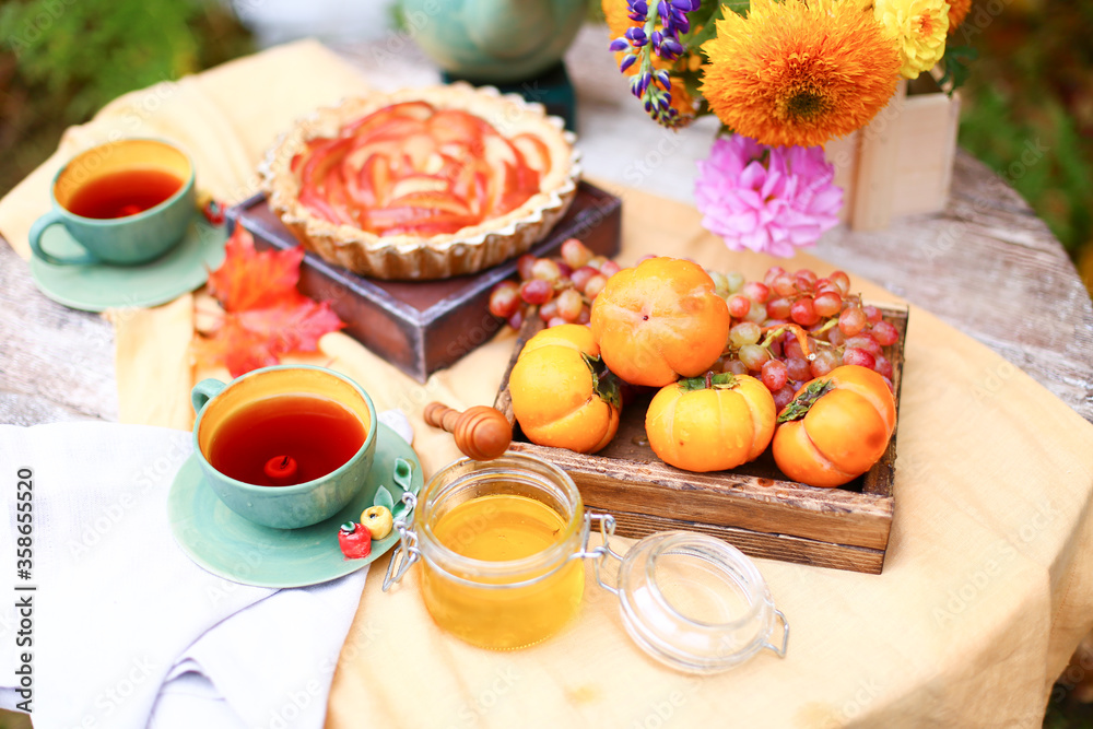 Autumn picnic. Tea party with beautiful kettle, cups at wooden table in garden. Harvest festival. Honey with stick, spoon, apple pie, persimmons, grapes, maple leaf, flowers, yellow linen tablecloth Autumn picnic. Tea party with beautiful kettle, cups at wooden table in garden. Harvest festival. Honey with stick, spoon, apple pie, persimmons, grapes, maple leaf, flowers, yellow linen tablecloth