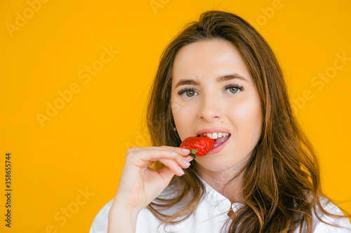 Beautiful girl is raising two strawberries to her cheeks. She is looking at the camera with temptation. Isolated on yellow background