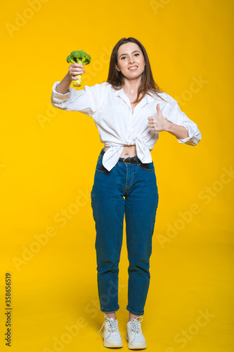 Portrait of a lovely pretty girl holding broccoli isolated over yellow background © ALEXSTUDIO