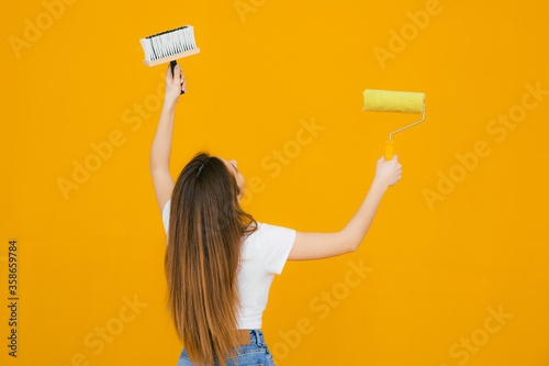 Young smiling beautiful woman in casual clothes holding paint roller for wall painting isolated on yellow background. Instruments, accessories, tools for renovation apartment room. Repair home concept