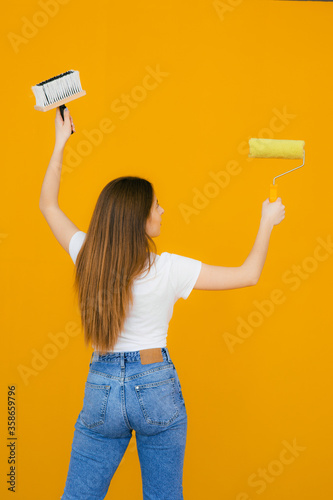 Young smiling beautiful woman in casual clothes holding paint roller for wall painting isolated on yellow background. Instruments, accessories, tools for renovation apartment room. Repair home concept