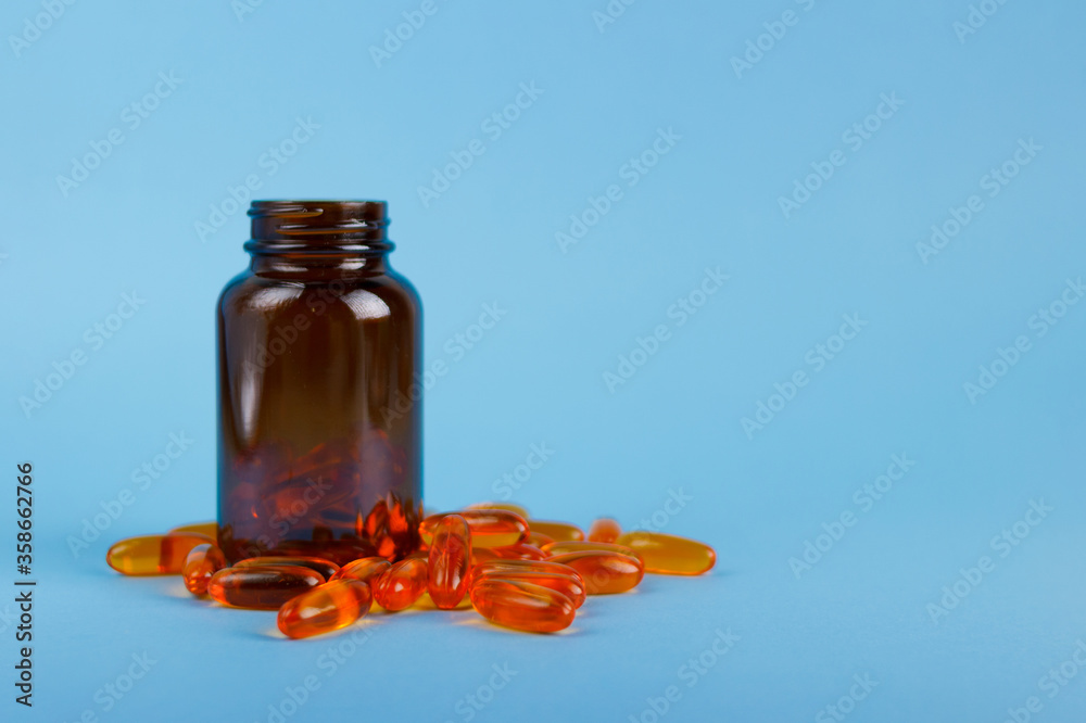 Close up orange fish oil capsules, omega 3, vitamin D in glass bottle jar isolated on blue background side view. Copy space for your design or text. Health care lifestyle concept. Close up orange fish oil capsules, omega 3, vitamin D in glass bottle jar isolated on blue background side view. Copy space for your design or text. Health care lifestyle concept.