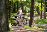 A white slender tall woman of sports physique in shorts and a T-shirt stands with a bicycle among the trees on the park path.