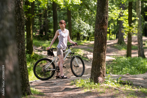 A white slender tall woman of sports physique in shorts and a T-shirt stands with a bicycle among the trees on the park path. © olga