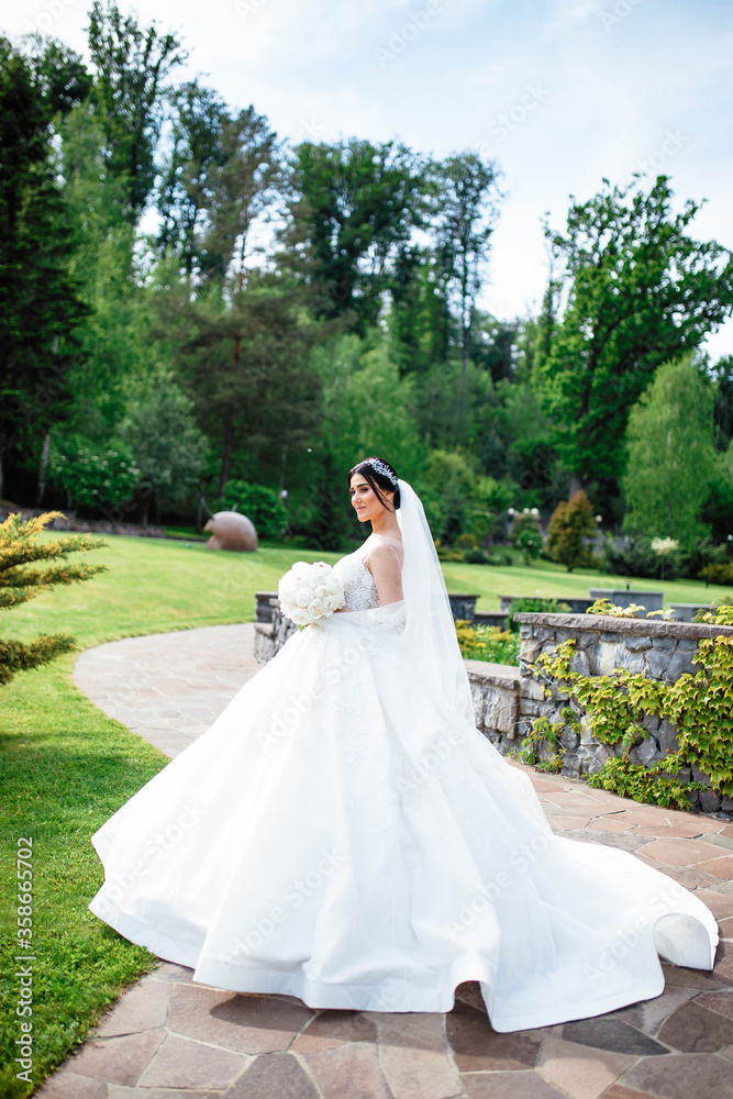 Young beautiful bride is standing on a garden. Beautiful bride in white wedding dress with bouquet in her hands the wind blows the veil. Sunshine portrait of happy bride outdoor in nature.