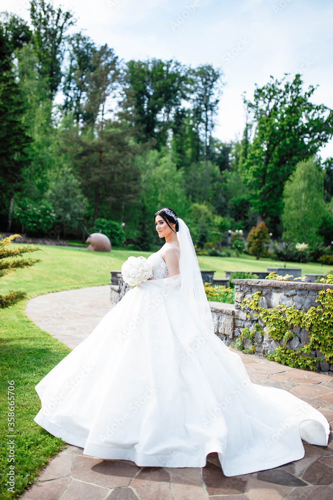 Young beautiful bride is standing on a garden. Beautiful bride in white wedding dress with bouquet in her hands the wind blows the veil. Sunshine portrait of happy bride outdoor in nature. Young beautiful bride is standing on a garden. Beautiful bride in white wedding dress with bouquet in her hands the wind blows the veil. Sunshine portrait of happy bride outdoor in nature.