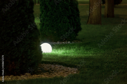 illumination backyard light garden with lantern electric lamp with a round diffuser in the green grass with thuja bushes in a park with landscaping, closeup night scene nobody.