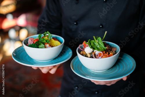 Male chef in a black tunic holding plates with dishes. Fresh salads with beans, cheese, cucumbers and herbs. The concept of fast preparing healthy, tasty and hearty meals.