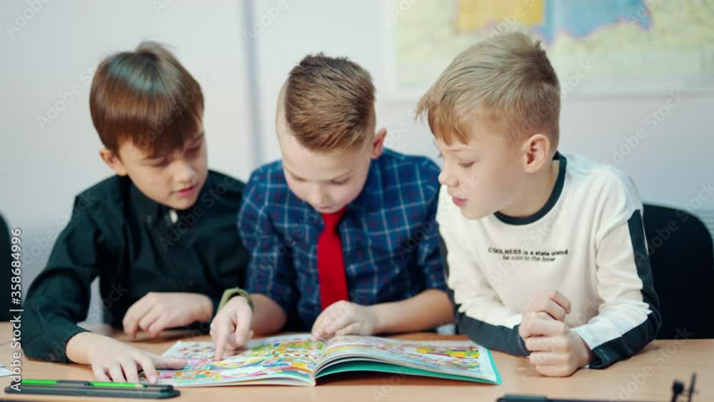Children reading a book. Schoolboys sitting together and looking into interesting book with pictures. Cute boy looking on camera and speak.