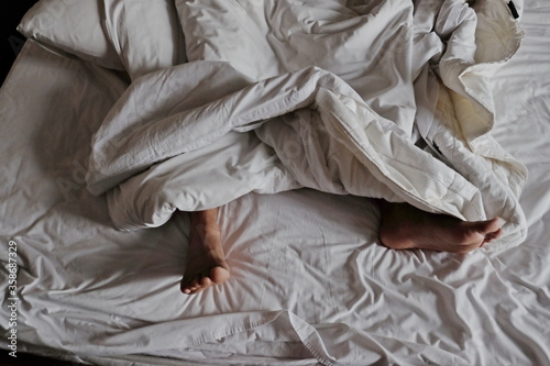 Close-up man feet alone in white bed, view of feet man sleeping and lying under white blanket and white bed , couples feet crossed under the duvet in bed
