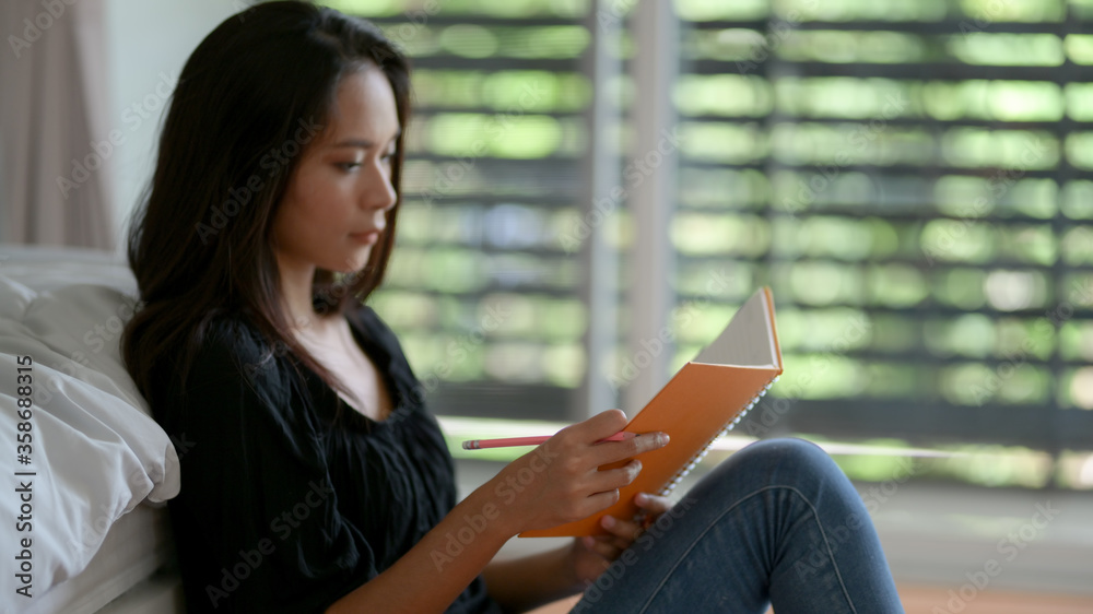 Fototapeta premium Female freelancer looking on her schedule book while sitting on floor at bedroom
