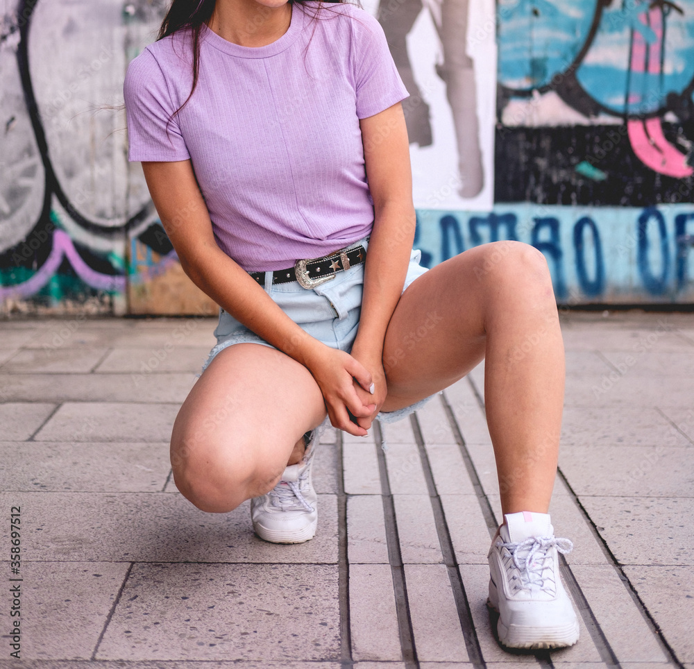 Sexy girl with bluejeans short, white sneakers, black belt with buckle and purple t-shirt with urban background
