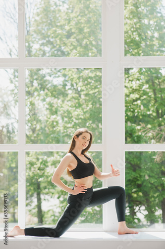 Beautiful woman in grey sportswear, bra and leggings practicing yoga, standing in anjaneyasana pose, girl doing Horse rider exercise, working out at home or in yoga studio with white walls